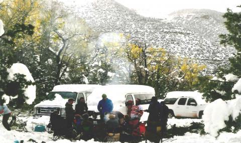 a group gathered around a campfire with snow coating the ground and vehicles parked behind them